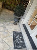 Full view showing metal urn with faux cedar and adjacent black rubber outdoor mat with scrollwork.