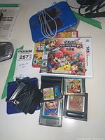 Blue Nintendo 2DS with grey AC adapter and pile of black flash cartridges, Super Smash Bros. and New Super Mario Bros. 2 cases visible