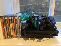 Front view of PlayStation 2 console, 4 wired controllers coiled on top, 10 PS2 game cases arranged to the side.