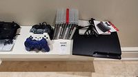 Photo showing PS3 console, four controllers in black, white, and blue, and 10 game cases.