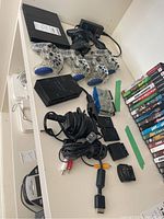Sony PlayStation 2 console accessories including five controllers, multiple game cartridges, cables, adapters, and power supplies spread out on a white shelf.