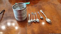 Eight glass and sterling silver coasters stacked, and five sterling silver flatware items (fork, coffee spoon, teaspoon, and three collector spoons) laid out on a wooden table.