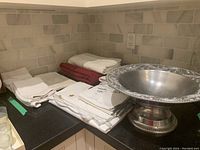Overview image showing stacked white hand towels, red tablecloth, white napkins with some embroidered pieces, and a silver metal bowl with a beaded edge.