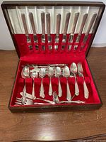 Wide view of the open wooden box showing multiple knives in the top section and spoons, forks, serving utensils in the bottom section with red velvet inserts.