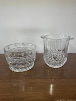 Front view of both crystal items side by side on a polished brown surface against a white wall showing details of cuts and shape.