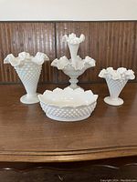 Full lot of four Fenton hobnail milk glass pieces: three different sized vases and one bowl, all white opaque glass with hobnail texture and ruffled edges, displayed on wooden surface.