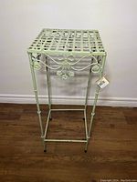 Full front view of light green metal vintage-look plant stand on wood floor against white wall.