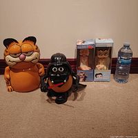 Front view showing four items: large Garfield ceramic coin bank, black Darth Vader Mr. Potato Head figure, two boxed bobbleheads (Marilyn Monroe and Betty Boop angel) with price tags visible, and a water bottle for scale.