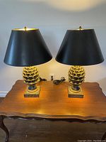 Front view photo showing both lamps with lit bulbs highlighting the pineapple bases and black shades on wooden table.