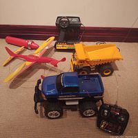 Lot photo showing the three toys: Caterpillar dump truck with corded remote, blue Chevy Silverado RC truck, and vintage corded plane on carpeted floor.