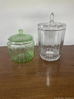 Front view of medium-sized green depression glass cookie jar with lid beside large clear crystal cookie jar with lid on a wooden surface.