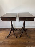 Front view of two vintage wooden end tables with rectangular tops, single drawer with brass pull, tripod pedestal bases, and claw feet with brass caps.