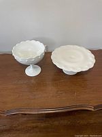 Front view of white Indiana milk glass tear drop pedestal fruit bowl and round pedestal cake stand with scalloped edge on wooden surface.