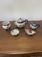 Front view of the full porcelain set on a wooden surface showing the teapot, creamer, sugar bowl, bonbon dish, and vase all with cobalt blue accents and floral painting.