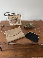 Four vintage purses including floral tapestry handbag, woven leather Holt Renfrew clutch, beaded black clutch, and floral metal clutch on wooden table.