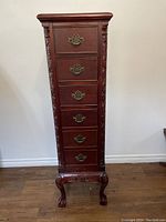 Front view of narrow mahogany-colored wooden cabinet with six drawers and ornate brass handles. Visible carved accents on legs and sides.