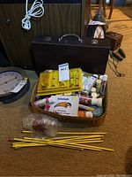 Photo of the box containing brown carrying case, assorted paint tubes, yellow knitting needles tangled in front, and yellow plastic case.