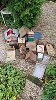 Overview photo showing variety of wooden bird houses and one metal bird feeder arranged on gravel outdoors near plants