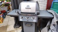 Close-up frontal view of Broil King propane BBQ grill with stainless steel lid and three control knobs, showing part of the cover and two metal grilling tongs on left side shelf.