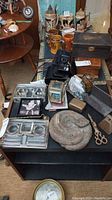 Photo of various collectible items including cast snake décor piece, wick trimming scissors, coasters, bell, and wooden box on a black table surface.