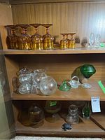 Wide photo of multiple shelves containing amber pressed glass stemware, clear glass bowls, green glass figurine, and salt and pepper shakers with one lid missing.