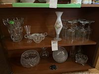 Shelf display with various glass items including a tall frosted vase with floral design, six Christmas themed wine glasses with green and red holly detailing, multiple small clear glass dishes, bowls, and a creamer.