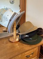 Wooden hat stand with multiple hats including beige felt with feather decoration, dark green felt hat with brush, and light woven straw hat.