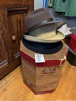 Three felt hats stacked on top of a vintage Stetson cardboard hat box showing wear and damage, on wood floor near wooden furniture.