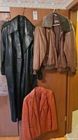 Three leather outerwear items hanging on a door: a black full leather coat, a brown leather bomber jacket with shearling collar, and a rust-colored leather blazer.