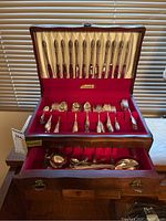 Set of silver plate flatware displayed in open wooden two-level box showing knives, forks, and spoons arranged neatly with red velvet and cream satin lining.