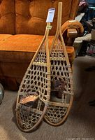Pair of traditional wooden snowshoes leaning against orange couch on carpeted floor, showing full length and bindings.