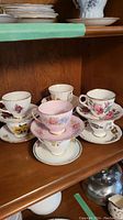 Eight vintage teacup and saucer sets displayed on shelf, varying floral designs and pastel colors.