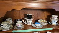 Image showing 11 teacups and saucers of various styles on wooden surface with different floral and colored designs, including demitasse sizes.