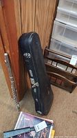 Violin case standing upright on carpeted floor next to a metal flute and magazines