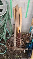 Photo of multiple long-handled yard tools (axes, pickaxe, sledge hammer) leaning against a concrete wall beside garden hose.