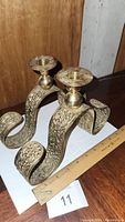 Set of two large brass tabletop candlesticks shown from an angled front-left side, highlighting engraved leaf and floral patterns and curled base.