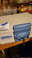 Photo of the white Brinks fireproof locking case focusing on the blue locking latch with a key inserted and the Brinks Home Security logo visible on the side.