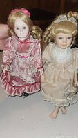 Two porcelain dolls, one in pink satin dress, one in beige lace dress, showing full front view and details of the dolls' faces and clothing.