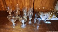 Photo showing full assortment of matching clear crystal stemware glasses in different styles and sizes, clear glass pedestal bowl, large glass measuring cup, and amber-tinted vintage glasses.