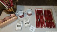 Photo of red tapered candles bundled in plastic trays, white emergency candles in their packaging, three small painted terra cotta planters, one large bucket terra cotta planter, and two coffee cups placed on a flat surface.