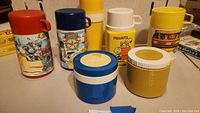 Five character thermoses and two insulated jars arranged on a table including Peanuts, Transformers, Aladdin, Galaxy Rangers, and Tung-I School Bus designs.