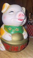 Front view of ceramic piggy bank showing details of face, green scarf with floral design, and gold coin with red Chinese characters.
