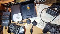 Sony PlayStation 2 slim console with open disc tray, two wired controllers, cables, and manuals arranged around it.