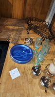 Four clear glass canisters with silver-tone lids, silver plated rose design jewelry box, blue plastic picnic plates with forks, clear vintage glass drinking cups, two black wire baskets on wood table