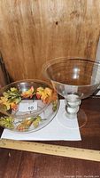 Clear glass floater bowl vase filled with artificial autumn colored leaves and berries next to oversized glass martini glass vase, measuring tape for scale.