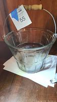 Front angled view of clear glass ice bucket shaped like a wooden apple basket, with vertical ribbed pattern and a metal frame base. Wooden handle attached with metal hardware.