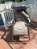 Full view of anti gravity lounge chair on wooden deck under daylight, showing padded fabric, neck pillow, cup holder, and metal frame in weathered condition.