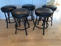 Five black swivel bar stools with round padded seats and black metal bases arranged together on a wood floor.