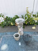 Photo of entire lot showing the two-piece cement birdbath, small cement plant pot, and black metal hanging plant holder placed on ground outdoors near plants.
