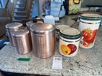 Vintage aluminum canisters with slight patina on surface, some toppers missing, placed beside three ceramic canisters with colorful vegetable illustrations (peppers, tomato).